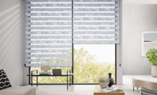 Modernes Wohnzimmer mit großen Fensterfronten und Doppelrollos mit Blumenmuster, lichtdurchlässig und blickdicht. Helle Einrichtung mit Ecksofa, Teppich, Couchtisch und Zimmerpflanze, minimalistisch und gemütlich. Blick ins Grüne durch bodentiefe Fenster – ideal für Interior Design und Wohnideen.