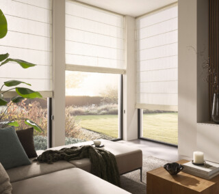 Lichtdurchflutetes, modernes Wohnzimmer mit Eckfensterfront, großem Sofa, Zimmerpflanze und transparenten, weißen LEHA Raffrollos Modell Cadenza.